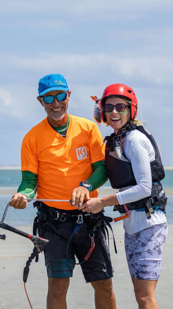 Moniteur OLK en lycra orange et élève avec casque rouge posant ensemble sur la plage après un cours de kitesurf, Pontal de Maceió