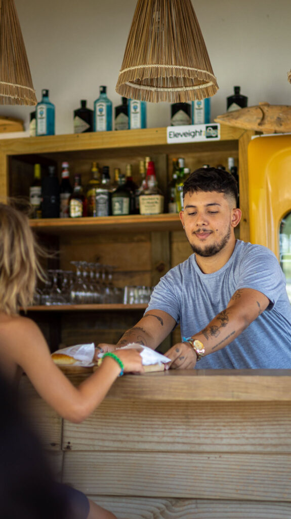 Barman servant un snack à une cliente au comptoir en bois du bar OLK House, Pontal de Maceió